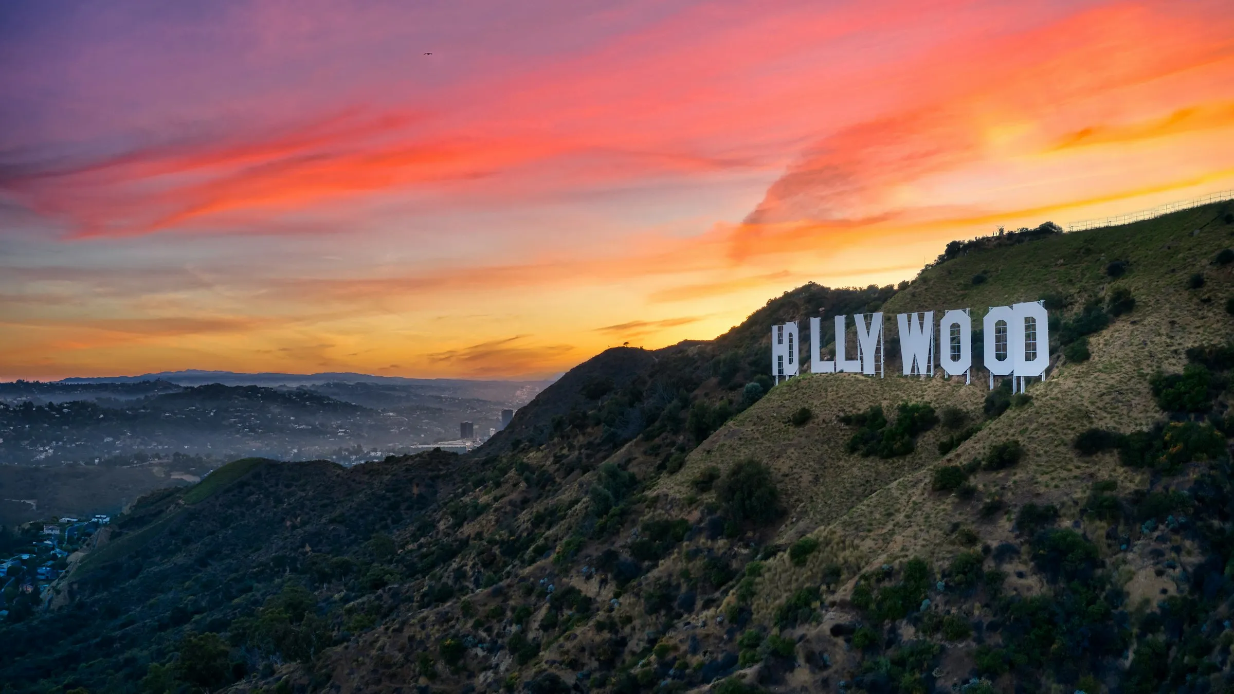 Letreiro de Hollywood na encosta do morro sob céu de pôr do sol rosa e laranja, com a cidade na névoa ao fundo